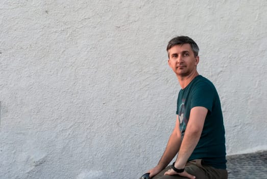 A middle-aged man sits against a white wall in summer clothes