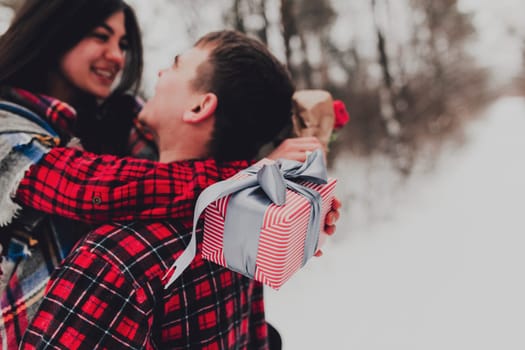 Boyfriend giving a gift box present with ribbon outdoor due to Saint Valentine day to her girlfriend. Snowy forest, trees with snow. Two are dressed in red clothes. Beautiful couple love each other