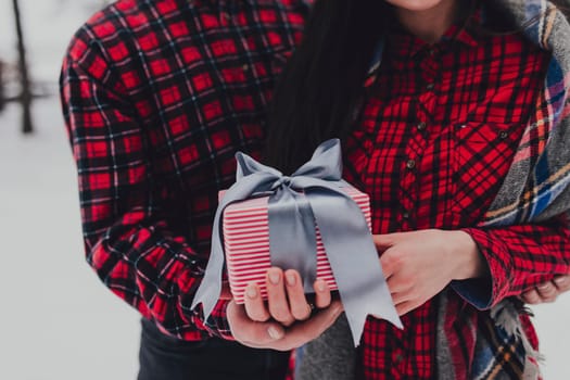 Boyfriend giving a gift box present with ribbon outdoor due to Saint Valentine day to her girlfriend. Snowy forest, trees with snow. Two are dressed in red clothes. Beautiful couple love each other