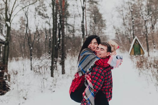 Boyfriend giving a gift box present with ribbon outdoor due to Saint Valentine day to her girlfriend. Snowy forest, trees with snow. Two are dressed in red clothes. Beautiful couple love each other