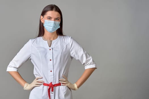 A young female doctor in a medical mask and a white coat stands on a gray background. Copy space. High quality photo