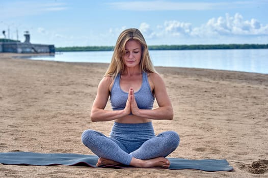 A woman sits in a lotus position on a sandy beach against the background of a river, yoga. High quality photo