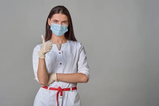 A doctor in a medical mask and a white coat shows his thumb on the background, copy space. High quality photo