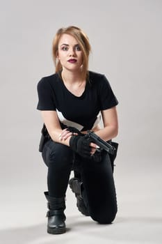 A young woman is on one knee and holding a gun in her hand on a gray background. High quality photo