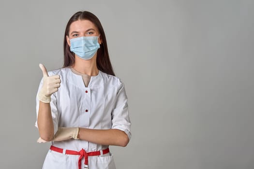 A doctor in a medical mask and a white coat shows his thumb on the background, copy space. High quality photo