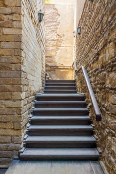 Architectural detail of outdoor wooden old weathered narrow staircase going up with wooden handrail, between two stone bricks walls, in abandoned building