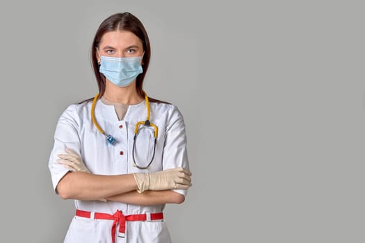 A young female doctor in a white coat with a stethoscope and a medical mask on a gray isolated background. Copy space. High quality photo
