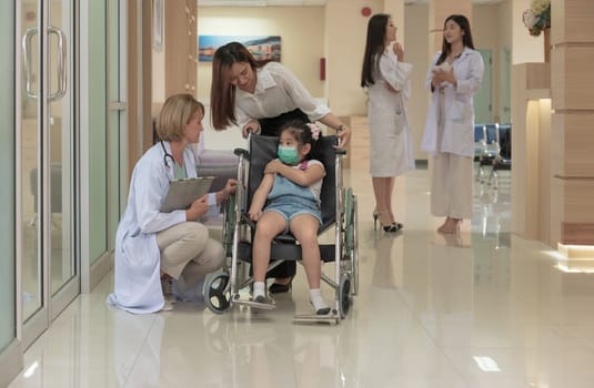 Little girl patient in wheelchair with medical facial mask and mother standing behind in hospital corridor with female doctor kneeling and check up. Medicine and healthcare services concept.