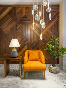 Modern orange armchair, lamp shade on small wooden table, planter with green bushes, and contemporary tall glass chandelier, in a hall with decorated wood cladding wall, and white marble floor