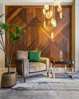Modern beige armchair, small wooden modern table, planter with green bushes, and contemporary tall glass chandelier, in a hall with decorated wood cladding wall, and white marble floor