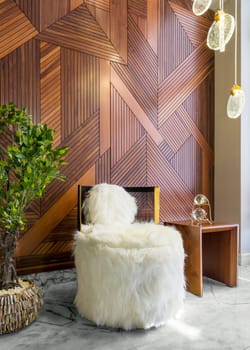 Modern white feather armless chair, small wooden modern table, and planter with green bushes, in a hall with decorated wood cladding wall, and white marble floor