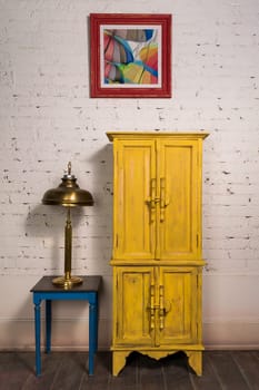 Still Life of vintage yellow cupboard, hanged framed painting, brass table lamp and vintage small table on dark wooden parquet floor and white bricks wall in Studio