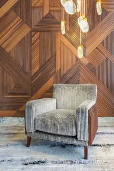 Modern grey armchair, and contemporary tall glass chandelier, in a hall with decorated wood cladding wall, and grey modern carpet