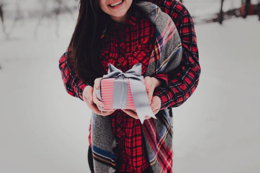 Boyfriend giving a gift box present with ribbon outdoor due to Saint Valentine day to her girlfriend. Snowy forest, trees with snow. Two are dressed in red clothes. Beautiful couple love each other
