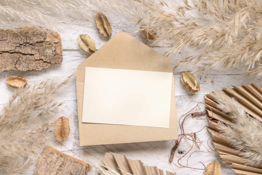 Blank card and envelope on white wooden table near dried plants, palm leaves and pampas grass top view. Boho scene with horizontal paper card flat lay. Greeting or Invitation card mockup, place for text