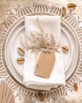 Wedding table place with blank gift tag attached to a napkin on bohemian placemat top view. Boho wedding label mockup Tropical beige template with dried plants, palm leaves and pampas grass, flat lay., place fot text
