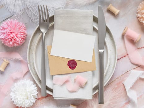 Wedding Table place with a paper card over envelope near flowers, silk ribbons and feathers top view. Romantic template with horizontal blank card on a plate flat lay. Card mockup, copy space