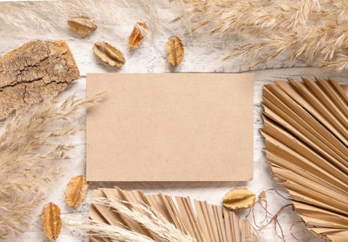 Blank card on white wooden table near dried plants, palm leaves and pampas grass top view. Boho scene with horizontal paper card flat lay. Greeting or Invitation card mockup, place for text