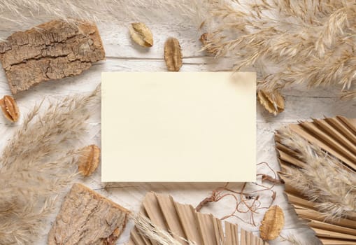 Blank card on white wooden table near dried plants, palm leaves and pampas grass top view. Boho scene with horizontal paper card flat lay. Greeting or Invitation card mockup, place for text