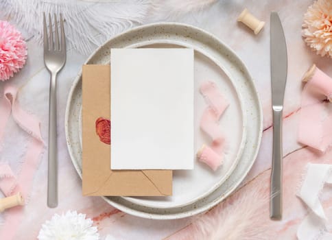 Wedding Table place with a paper card over sealed envelope near flowers, silk ribbons and feathers top view. Romantic template with horizontal blank card on a plate flat lay. Card mockup, copy space