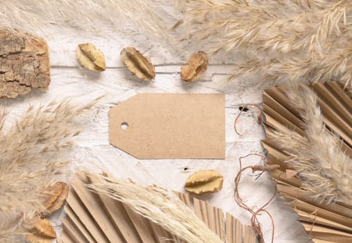 Blank gift tag on white wooden table near dried plants, palm leaves and pampas grass top view. Boho scene with horizontal paper label flat lay, place for text