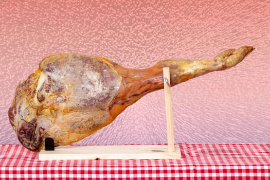 Spanish food. Closeup of a front leg of a Spanish Serrano ham on a wooden stand on a red checkered tablecloth over abstract pink cube block raster background. Template for your design.