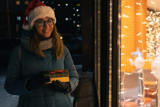 Pretty Woman in Santas hat with gifts box near illuminated cafe window. She waves her hand and waits for her beloved partner. Xmas presents holidays, or shopping on New Year or Christmas concept