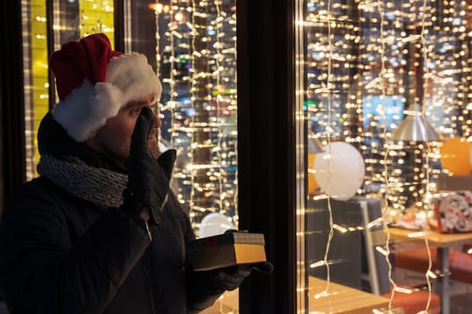 Man in Santas hat with gifts box near illuminated cafe window. He waves hand and waits for her beloved partner. Xmas holidays or New Year or Christmas concept