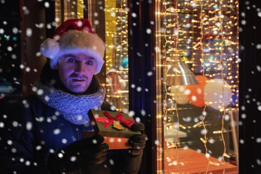 Man in Santas hat with gifts box near illuminated cafe window.