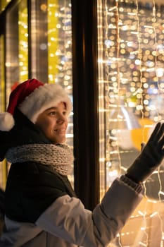 Boy in Santas hat looking and dreaming in illuminated shop window. Xmas presents holidays, or shopping on New Year or Christmas concept