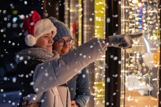 Pretty Woman and her son in santas hat looking and dreaming in illuminated shop window. Xmas presents holidays, or shopping on New Year or Christmas concept