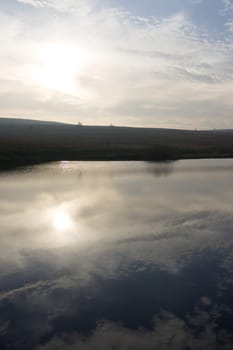 Vertical image of blue cloudy sky reflected in redmires top reservoir. Looking into the sun over heather covered peak district moor. Winter season.