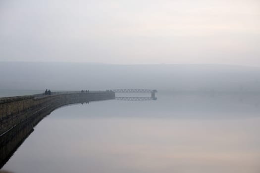 Sheffield, United Kingdom, 7th November, 2020: Bleak winter scene over redmires reservoir. Groups of people walk the dam wall. atmospheric fog or mist