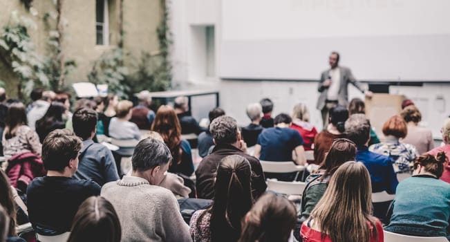 Male speaker giving presentation in lecture hall at university workshop. Audience in conference hall. Rear view of unrecognized participant. Scientific conference event. Copy space on whitescreen.