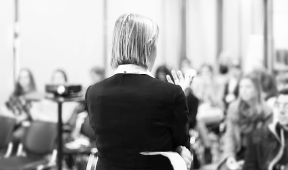 Female speaker giving a talk on corporate business conference. Unrecognizable people in audience at conference hall. Business and Entrepreneurship event.