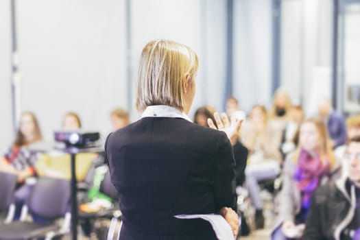 Female speaker giving a talk on corporate business conference. Unrecognizable people in audience at conference hall. Business and Entrepreneurship event.