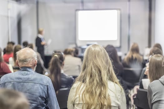 Business and entrepreneurship symposium. Female speaker giving a talk at business meeting. Audience in conference hall. Rear view of unrecognized participant in audience. Copy space on whitescreen.
