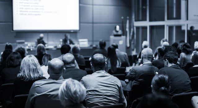 Business and entrepreneurship symposium. Speaker giving a talk at business meeting. Audience in the conference hall. Rear view of unrecognized participant in audience.