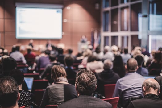 Business and entrepreneurship symposium. Speaker giving a talk at business meeting. Audience in the conference hall. Rear view of unrecognized participant in audience.