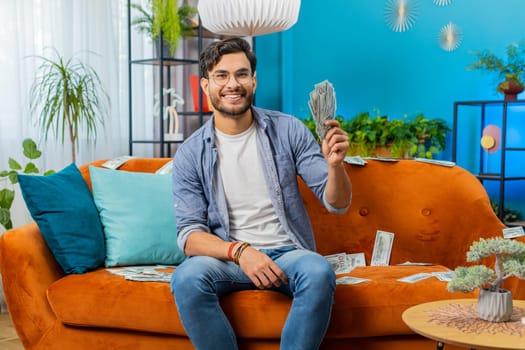Successful happy rich winner Indian Arabian man counting money cheering up with high profits, lottery game win, satisfied of wealth income earnings salary, on home couch full of dollar cash banknotes