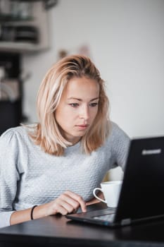 Female freelancer in her casual home clothing working remotly from her dining table in the morning. Home kitchen in the background