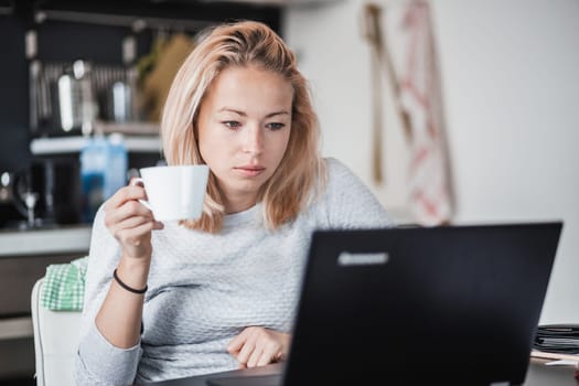 Female freelancer in her casual home clothing working remotly from her dining table in the morning. Home kitchen in the background
