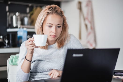 Female freelancer in her casual home clothing working remotly from her dining table in the morning. Home kitchen in the background