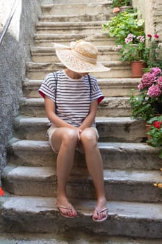 Beautiful female tourist wearing big straw sun hat and shorts sitting and relaxing on old stone house stairs during summer travel Mediterranean costal village on hot summer day.