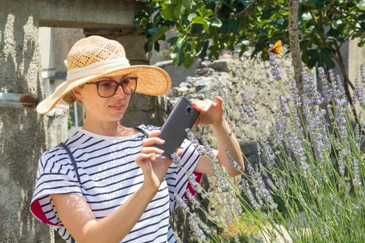 Beautiful blonde young female traveler wearing straw sun hat taking photo of lavander while enjoying summer vacation in an old traditional village on Mediterranean cost