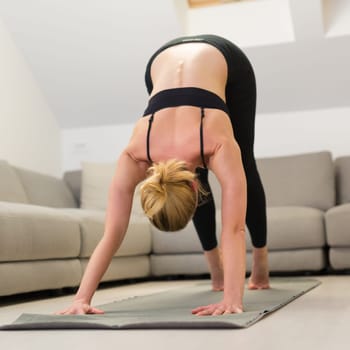 Beautiful blonde woman doing home workout indoors. Woman practice yoga at home. Fit girl using workout tutorials for healthy active lifestyle. Woman using quarantine for home workouts. Stretching