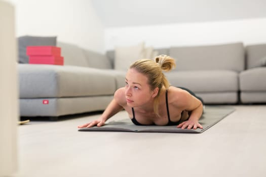 Beautiful blonde woman doing home workout indoors. Woman practice yoga at home. Fit girl using workout tutorials for healthy active lifestyle. Woman using quarantine for home workouts. Stretching