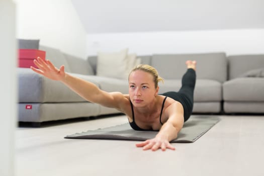 Beautiful blonde woman doing home workout indoors. Woman practice yoga at home. Fit girl using workout tutorials for healthy active lifestyle. Woman using quarantine for home workouts. Stretching