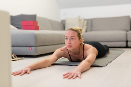 Beautiful blonde woman doing home workout indoors. Woman practice yoga at home. Fit girl using workout tutorials for healthy active lifestyle. Woman using quarantine for home workouts. Stretching
