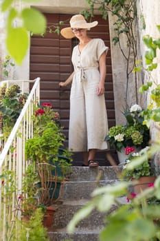 Beautiful female tourist wearing big straw sun hat and sundress standing on old vintage stone house staircase during summer travel on Mediterranean cost on hot summer day